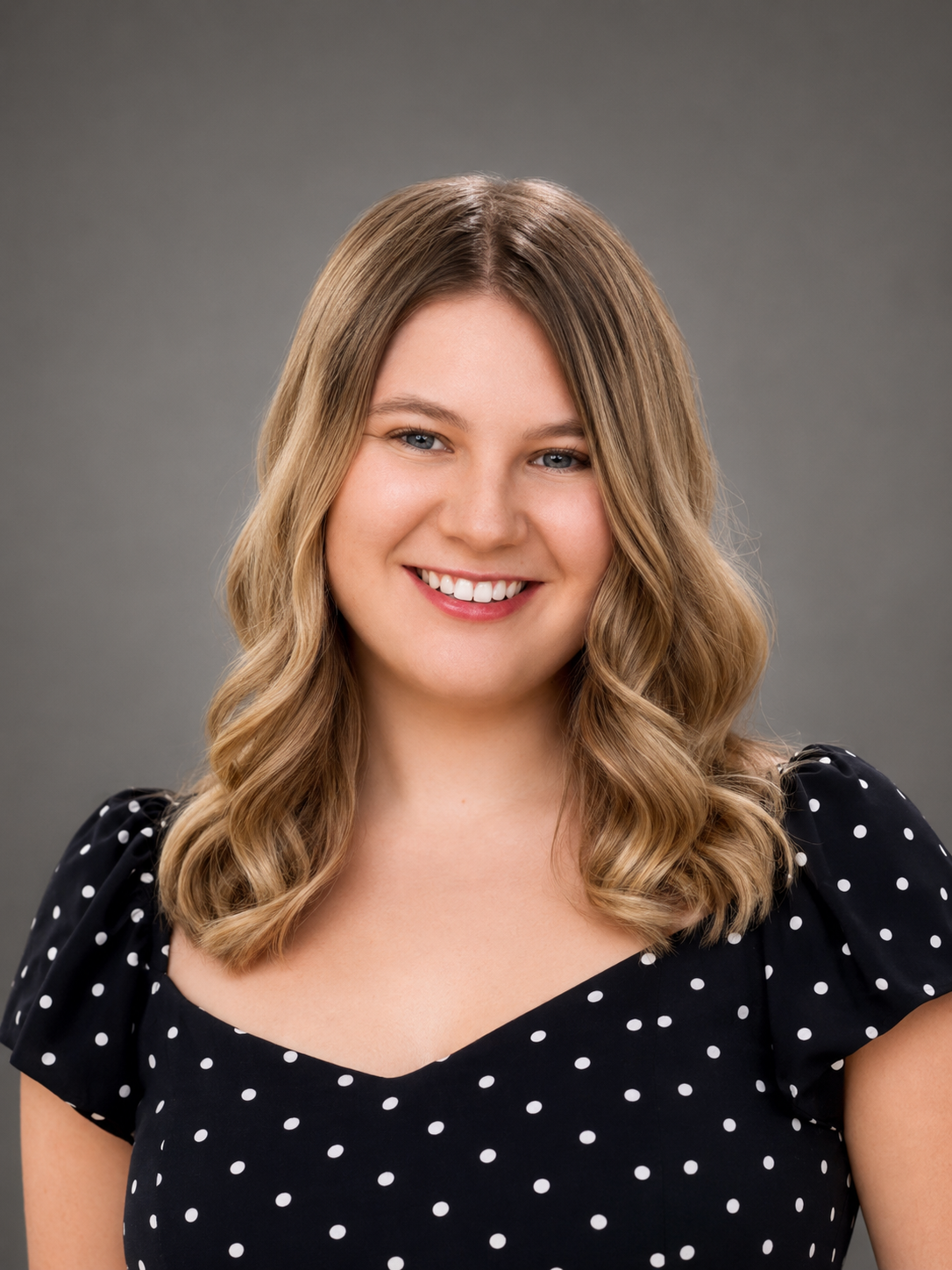 [NAME] smiles toward the camera in a professional head-and-shoulders portrait against a gray studio background. She is wearing a black dress with white polka dots and has shoulder-length wavy blonde hair.