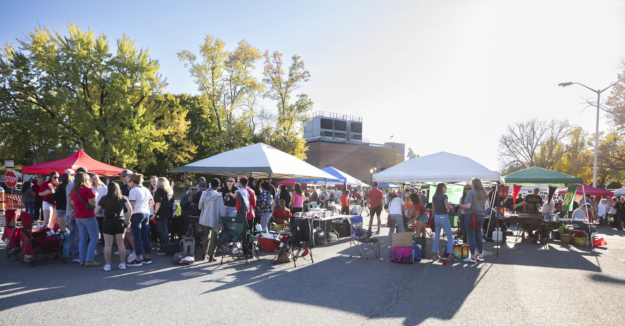 A vibrant tailgate at East Stroudsburg University with alumni and students socializing under tents. People are grilling food and enjoying the sunny fall weather with colorful autumn trees in the background.